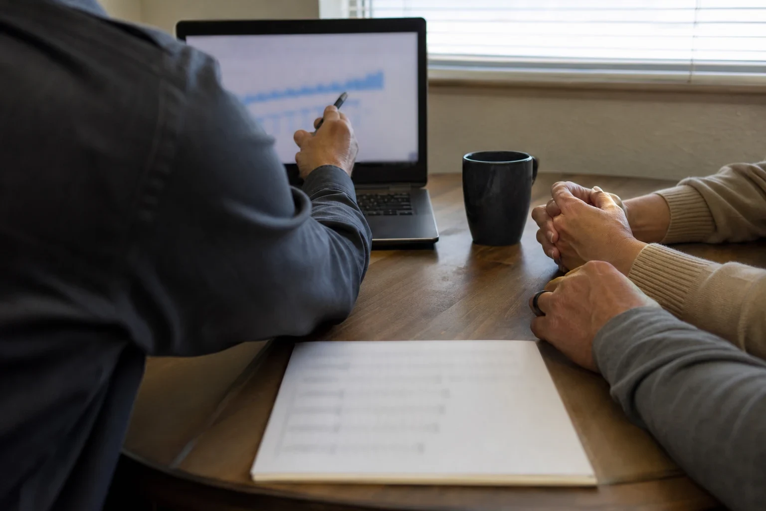 Agent seen from behind pointing a pen at a laptop chart while a couple's clasped hands rest on a wooden table beside a printed summary and a dark coffee mug
