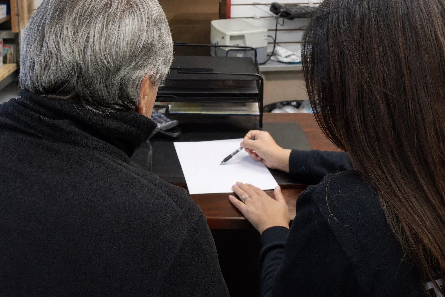 Over-the-shoulder view of a gray-haired man and a woman with a pen reviewing a sheet of paper at a desk in a small back office with a printer and phone