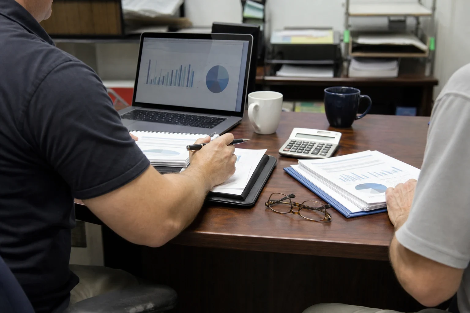 Two people at a wooden office desk reviewing printed charts, with a laptop showing graphs, a calculator, coffee mugs, reading glasses and a pen in hand
